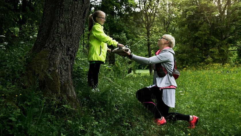 kobieta w stroju sportowym klęczy przed małą dziewczynką podając jej kije do nordic walking w tle las i góry
